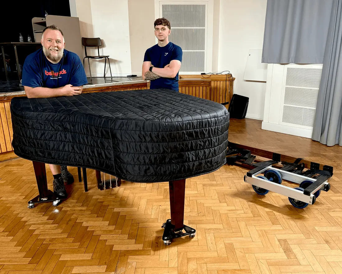 Two piano movers with a covered grand piano on a school hall floor, ready for transport