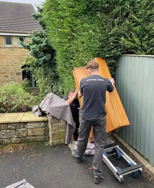 Piano movers carrying an upright piano on a trolley past a tight garden access