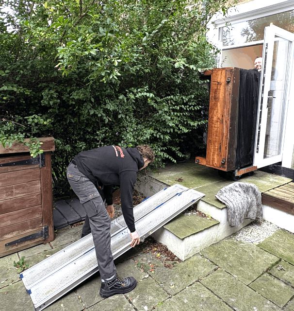Upright piano being loaded via ramp from a garden